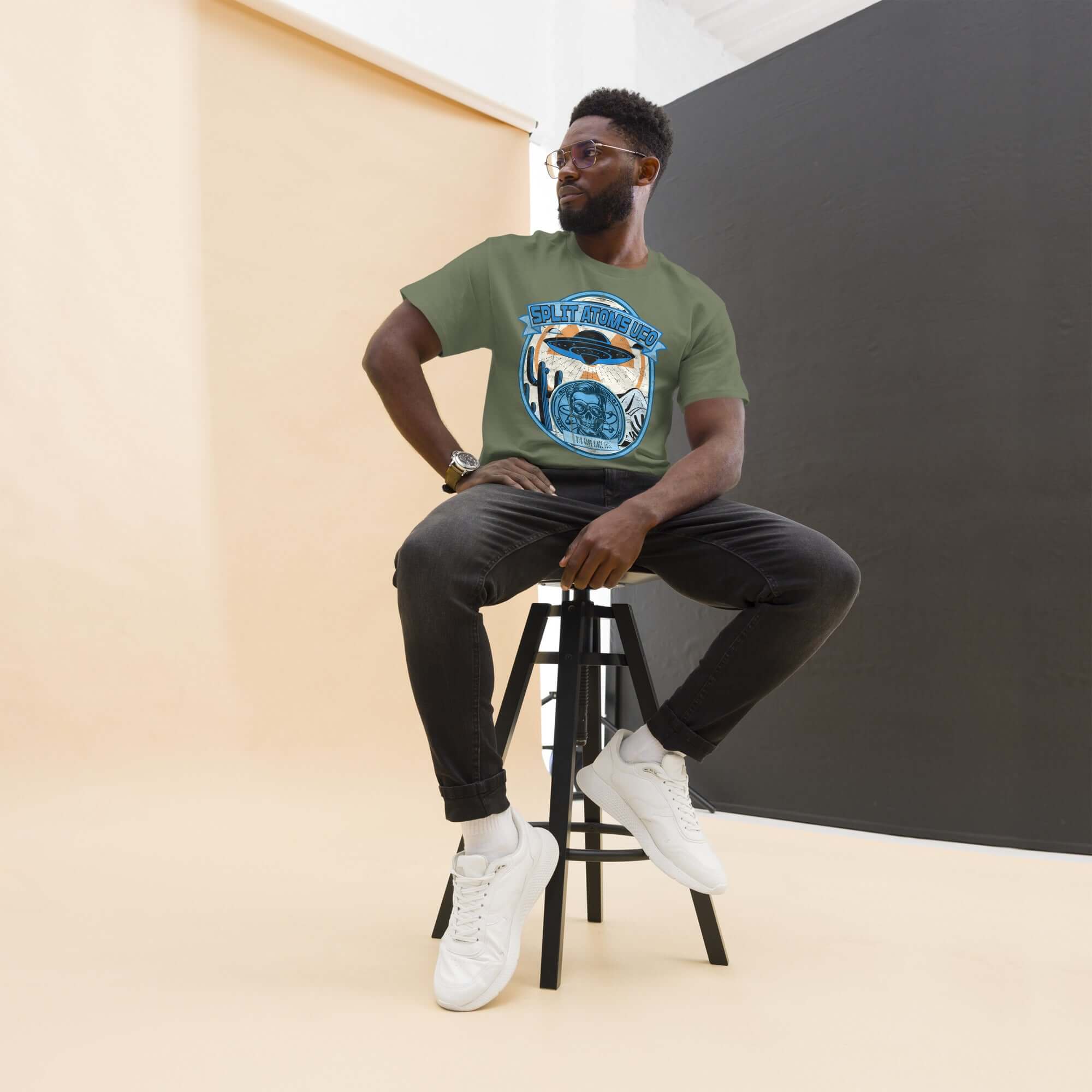 Model wearing Split Atoms UFO Gang T-Shirt with a retro alien desert design, sitting on a stool in a studio setting.