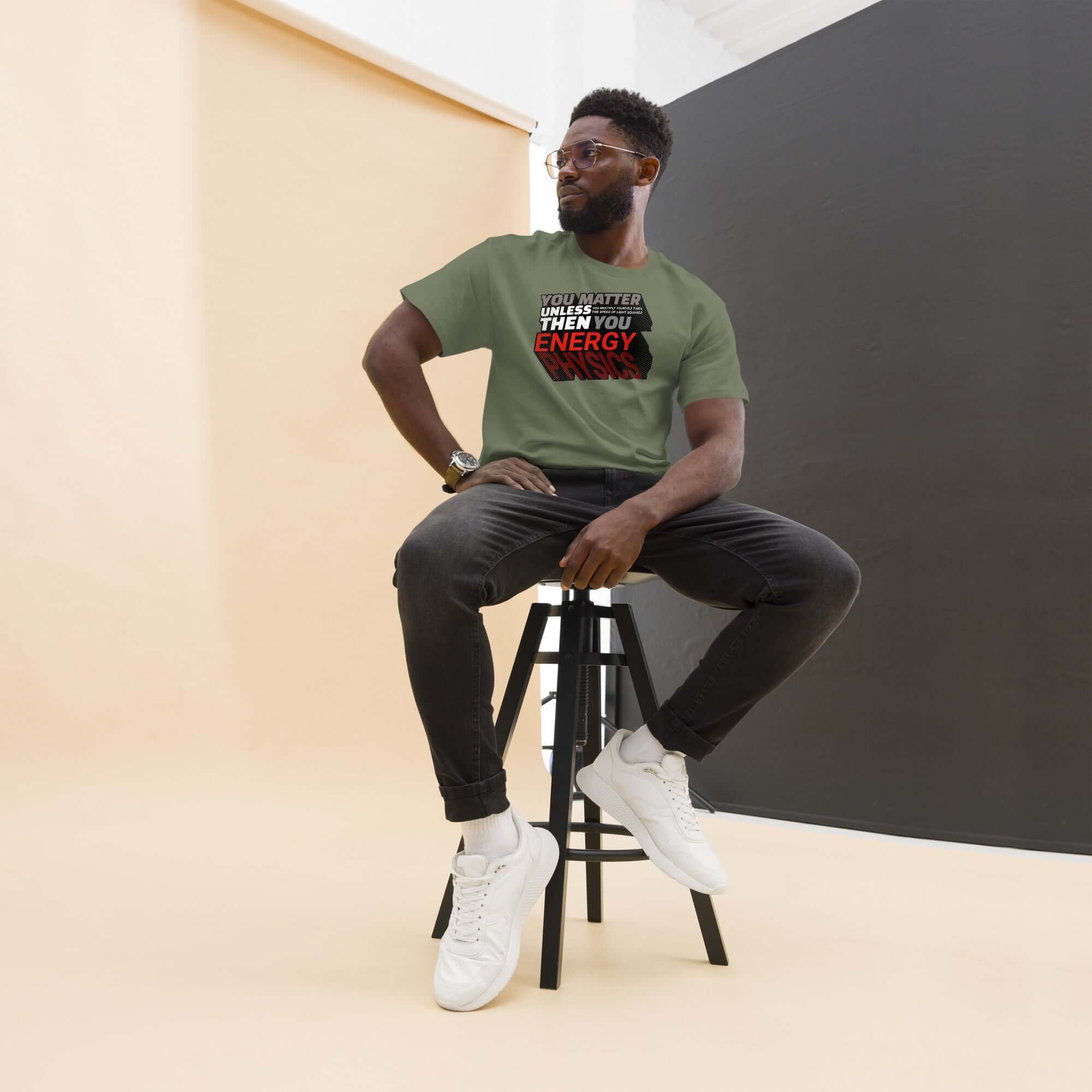 Man wearing Einstein's E=mc² equation humor t-shirt in green with bold text sitting on a stool in bright modern studio.