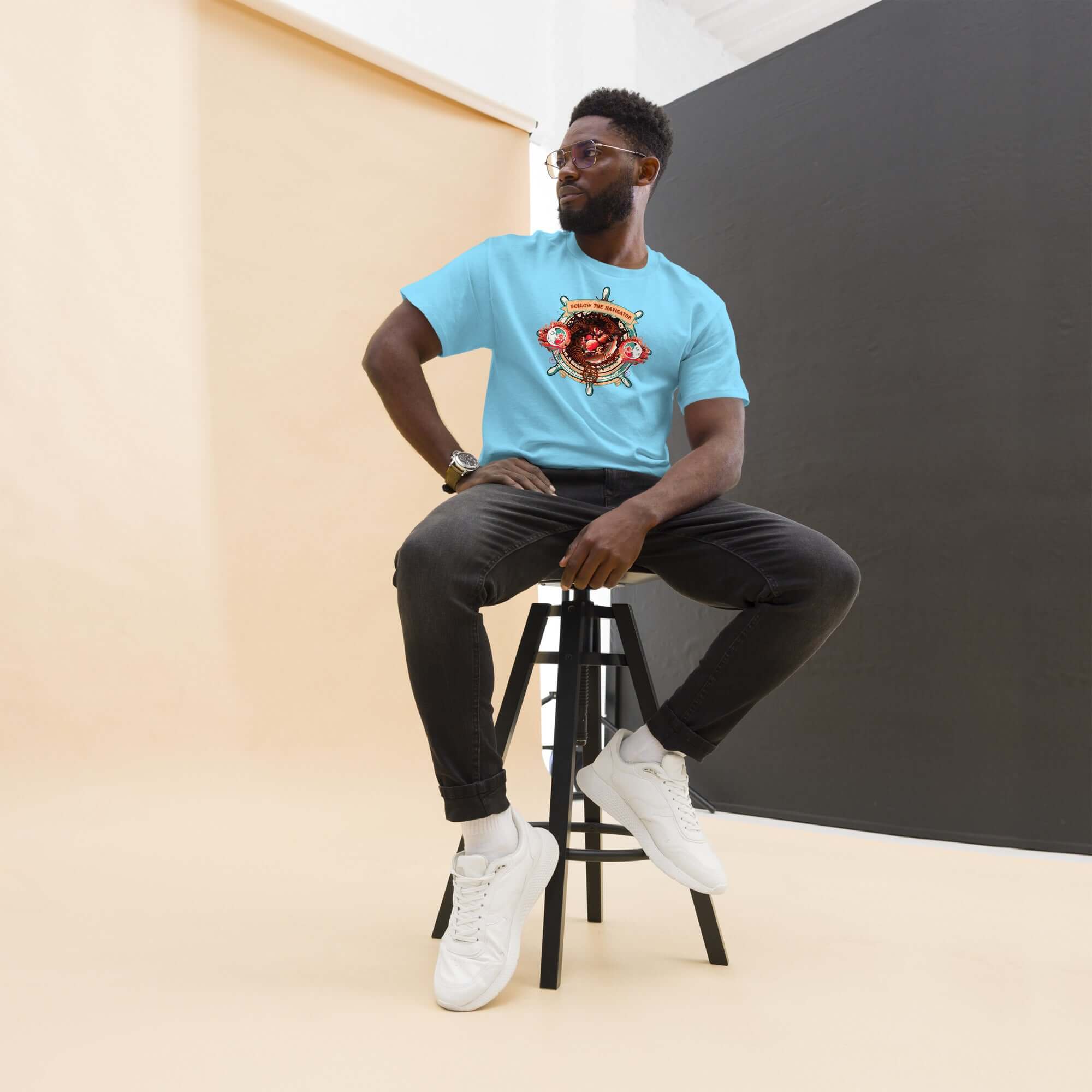 Model wearing Follow The Navigator T-Shirt, turquoise blue with cosmic compass design, sitting on stool in modern studio setting.