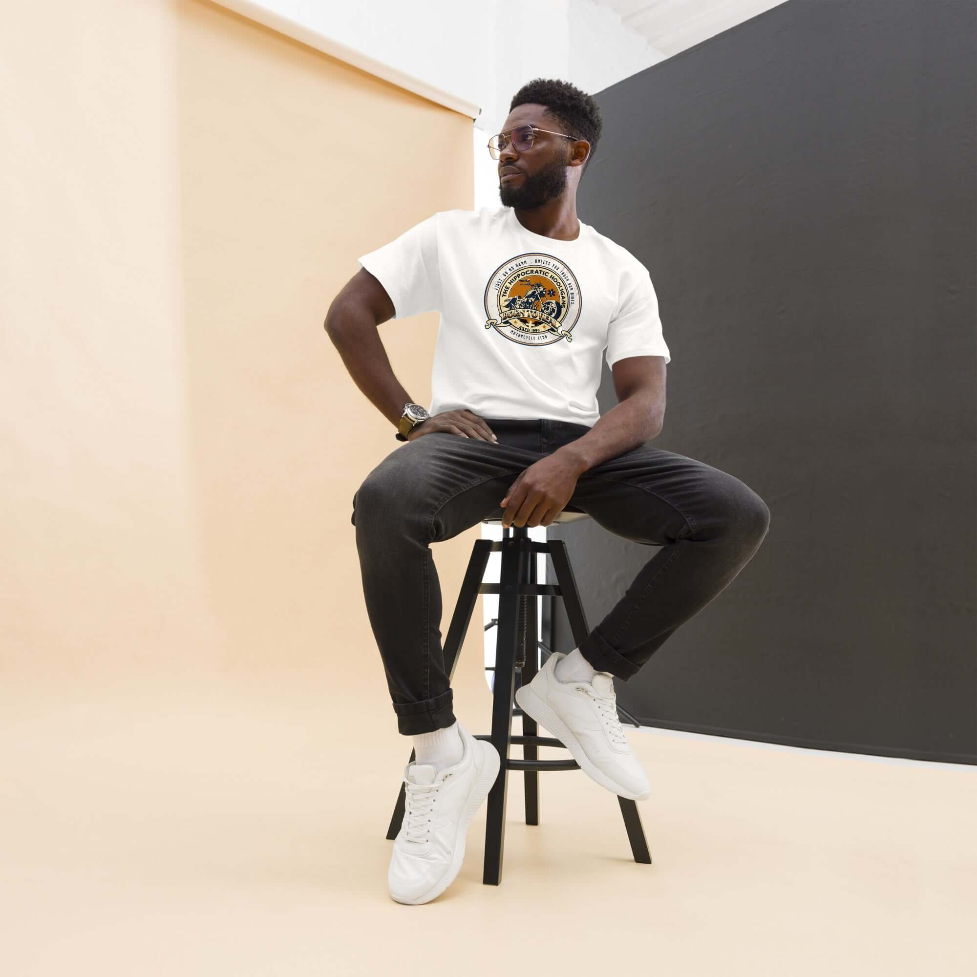 Man wearing a Hippocratic Hooligans Motorcycle Club T-Shirt, featuring a biker patch design, seated on a stool.